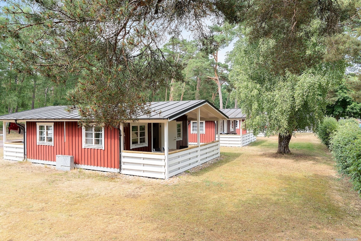 Uma cabana vermelha com varanda e uma pequena construção anexa, rodeada por árvores.