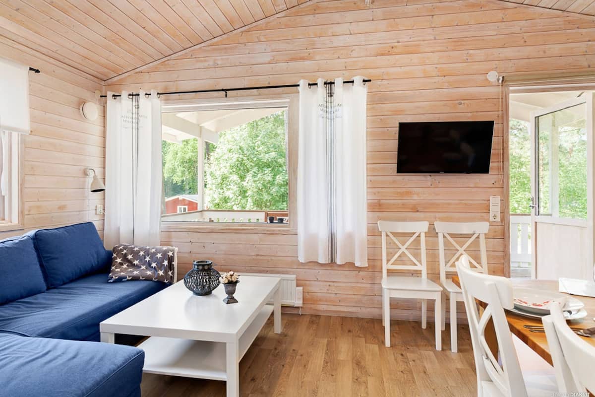Quarto com paredes de madeira, sofá azul, cadeiras brancas, mesa de jantar e televisão na parede.