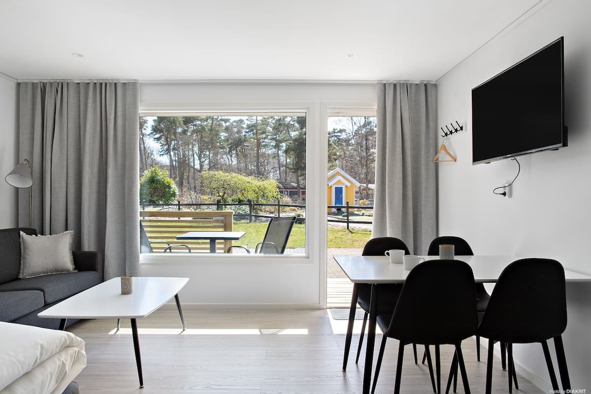 Uma sala de estar moderna com sofá, mesa de jantar com cadeiras, TV montada na parede e uma grande janela com vista para o exterior.