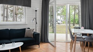 A modern living room with a dark-colored sofa, a white coffee table, and a dining table with chairs. There is a large window with curtains, a floor lamp, and a view of trees and a car outside.