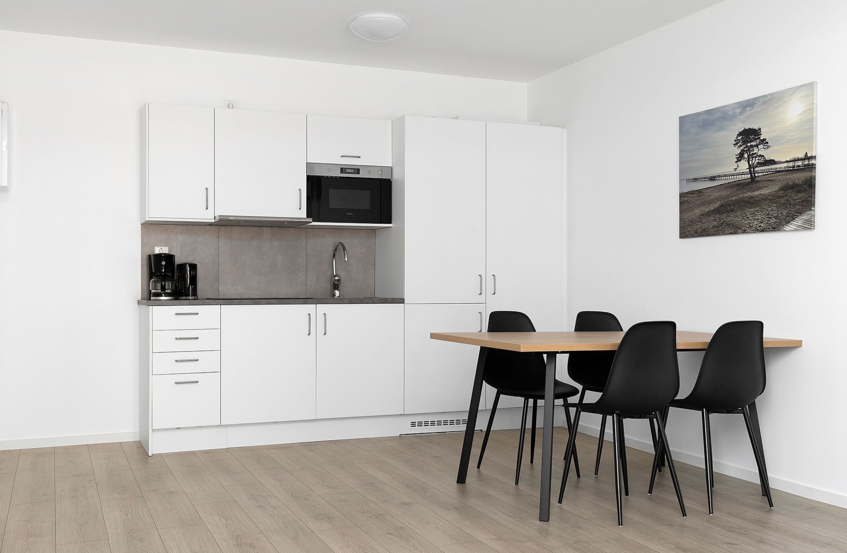 A modern kitchen with white cabinets, a microwave, and a coffee maker. There is a dining table with four chairs and a painting on the wall.