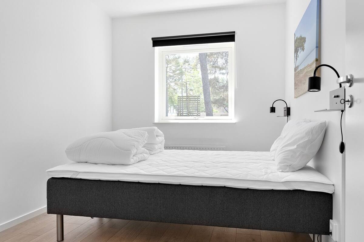 Une chambre à coucher minimaliste, avec un lit, une fenêtre donnant sur des arbres et une lampe fixée au mur.