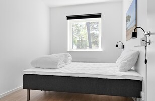 Une chambre à coucher minimaliste, avec un lit, une fenêtre donnant sur des arbres et une lampe fixée au mur.