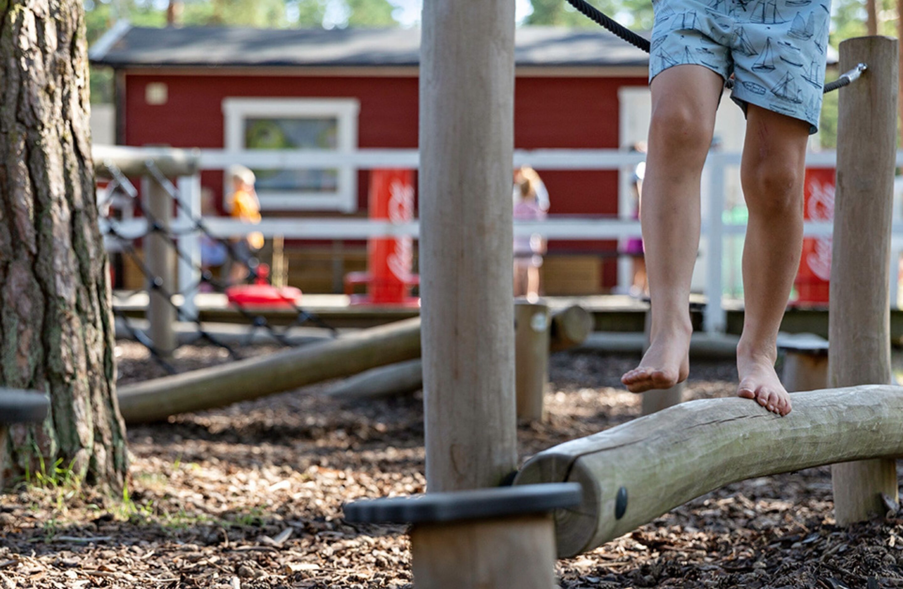 A child balancing on a log in an outdoor play area with wooden structures and a red building in the background.