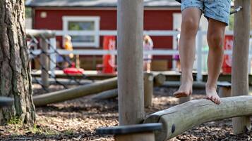 A child balancing on a log in an outdoor play area with wooden structures and a red building in the background.