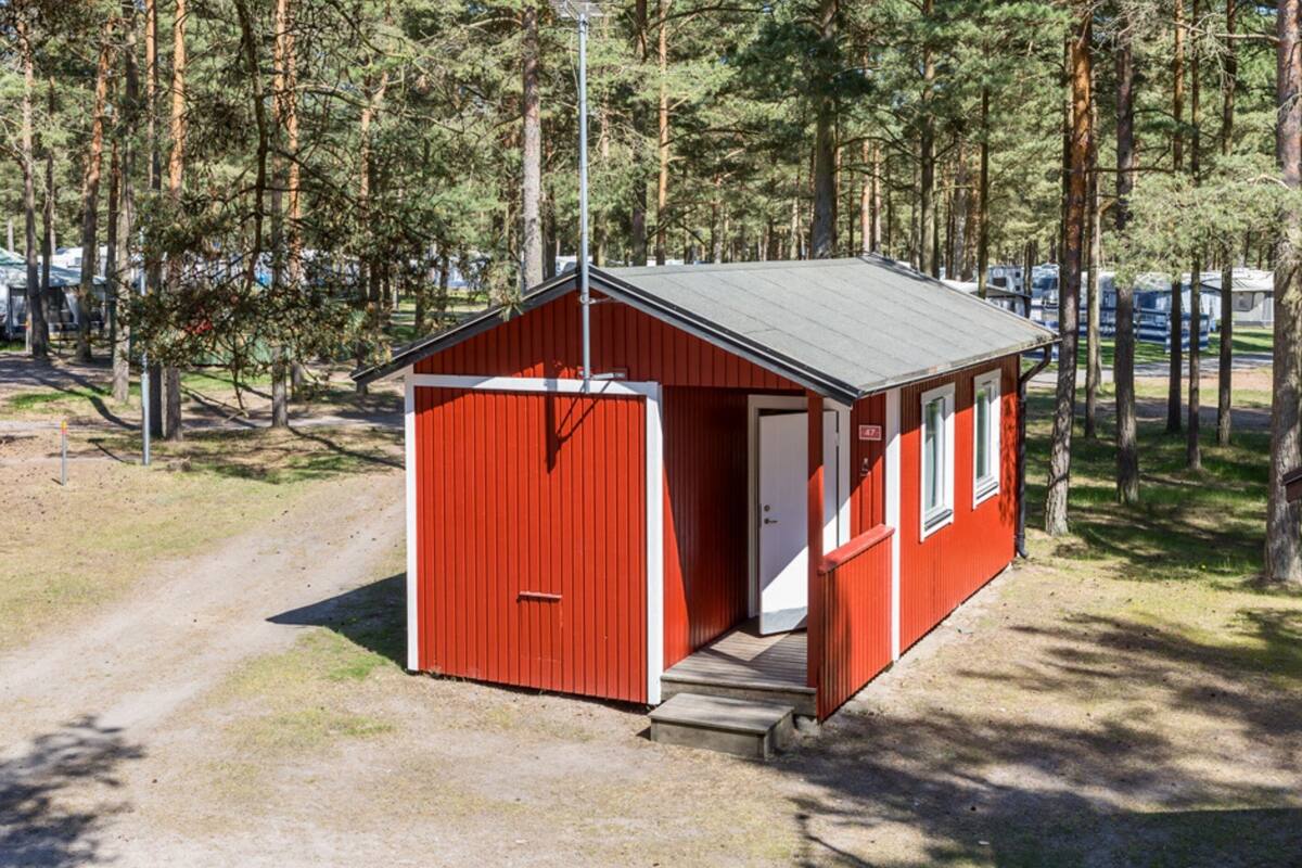 Une petite cabane rouge, dotée d’une porte et de fenêtres, entourée d’arbres et bordée d’un chemin de gravier.