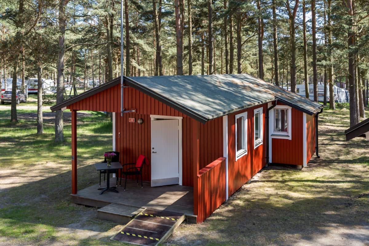 Une petite cabane à un seul étage, avec une façade rouge et un toit vert, située dans une zone boisée. Elle est dotée d’une terrasse dallée et d’une petite table entourée de chaises.