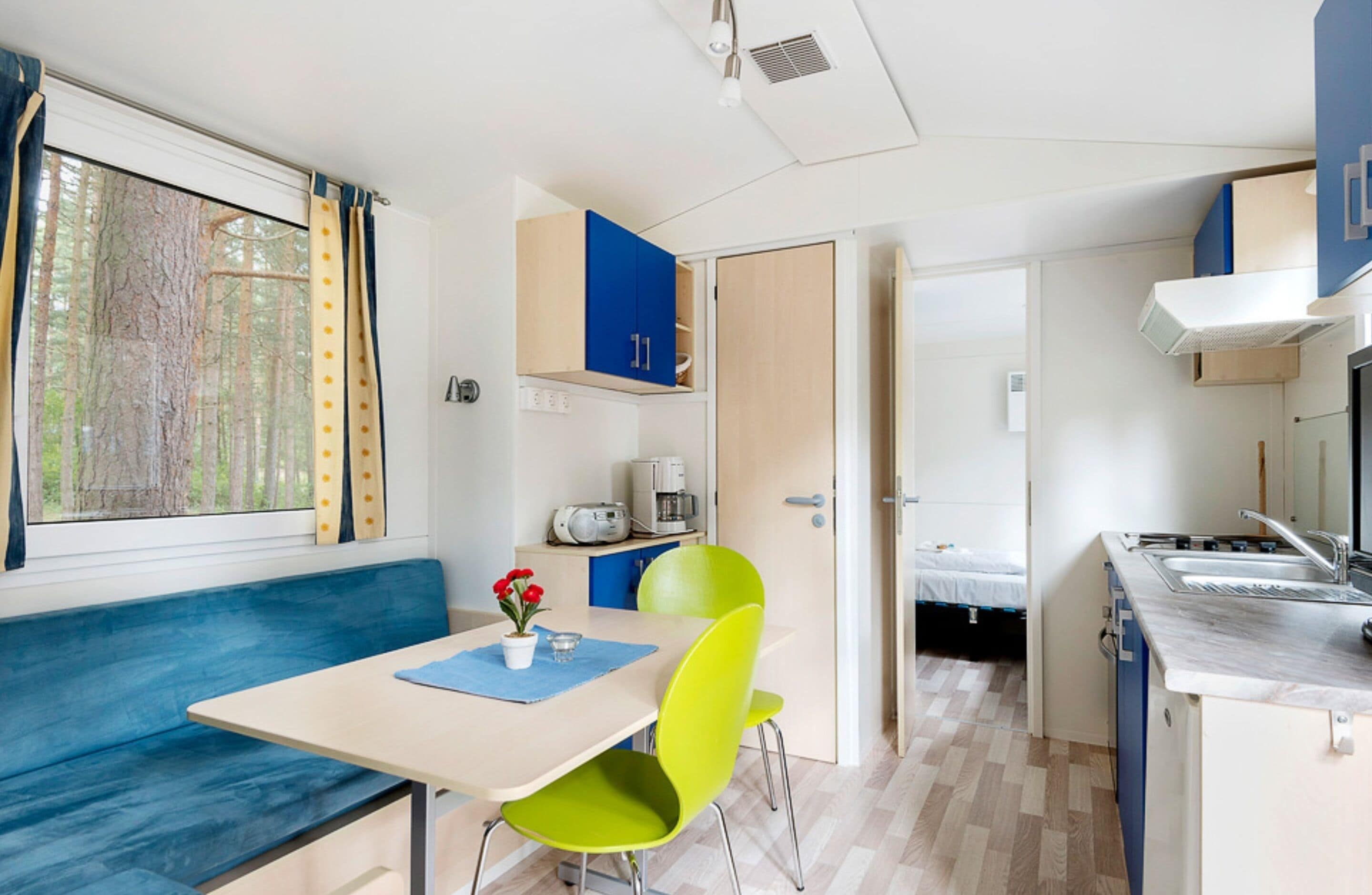A compact room with a dining table, bright green chairs, a bench, a kitchenette, and a bed.