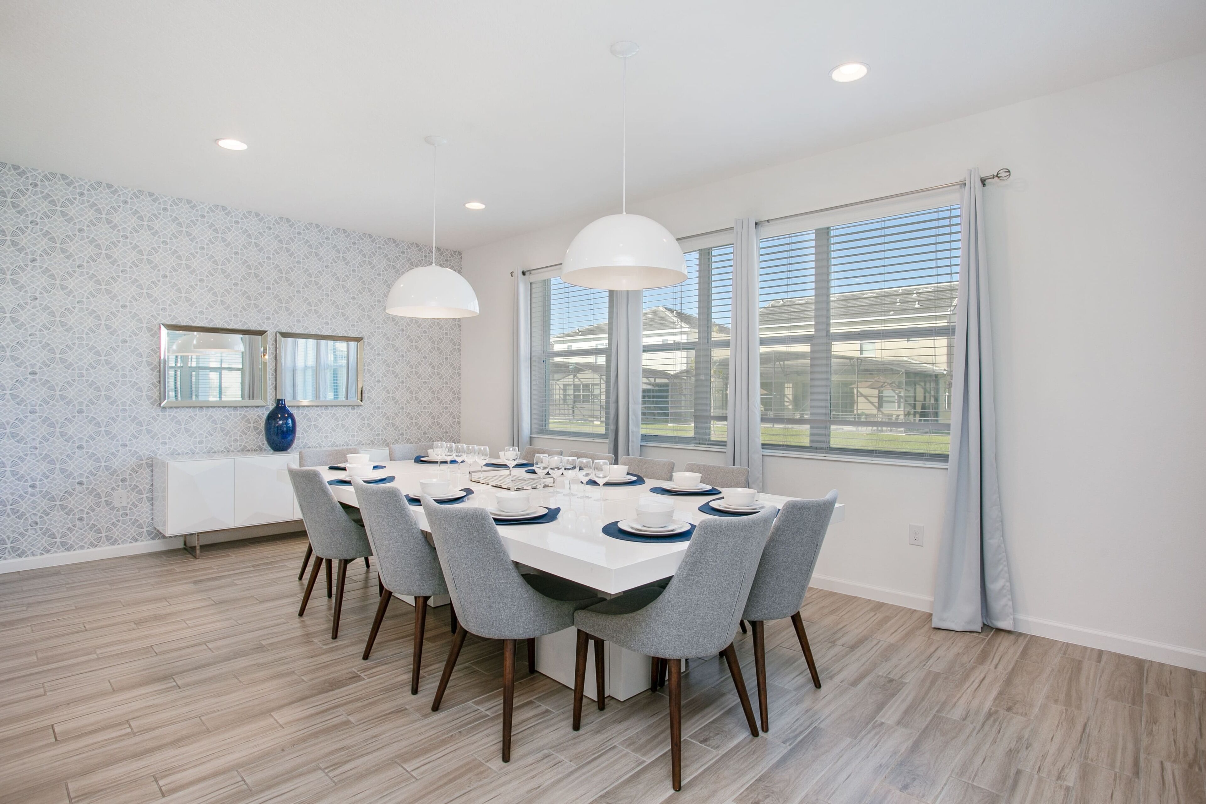 A dining area with a large table set for six, surrounded by grey chairs. There are two pendant lights above the table, and a window with blinds on the right.