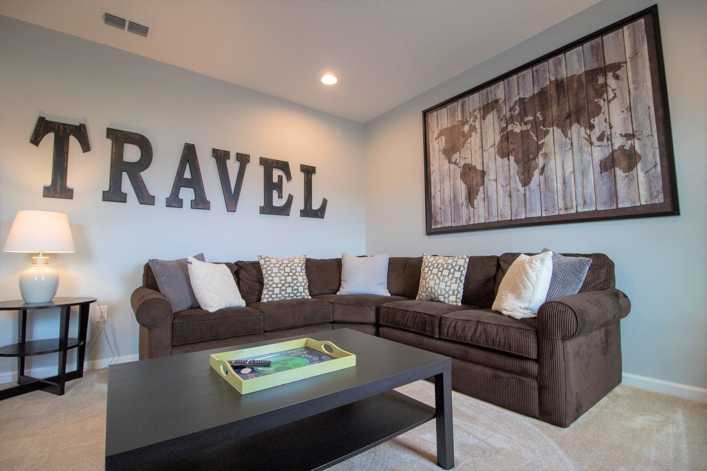 A living room with a brown sectional sofa, a coffee table, a world map artwork, and a 'TRAVEL' sign on the wall.