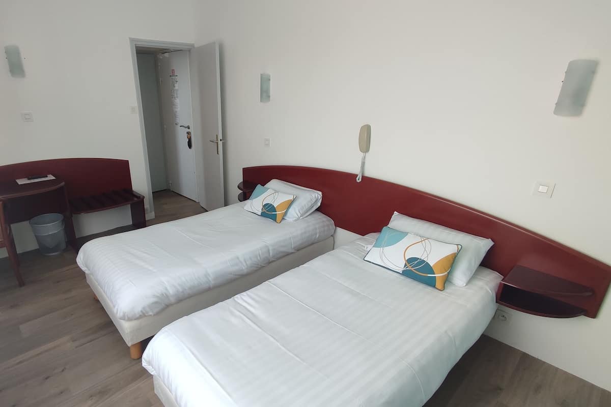 Ein Hotelzimmer mit zwei Einzelbetten, einem Schreibtisch mit Stuhl, einem kleinen Tisch und einer Mülleimer.
