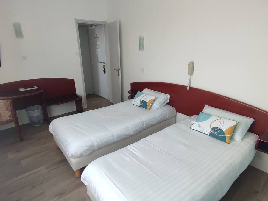 Habitación de hotel con dos camas individuales, un escritorio con silla, una mesita pequeña y un cesto de basura.