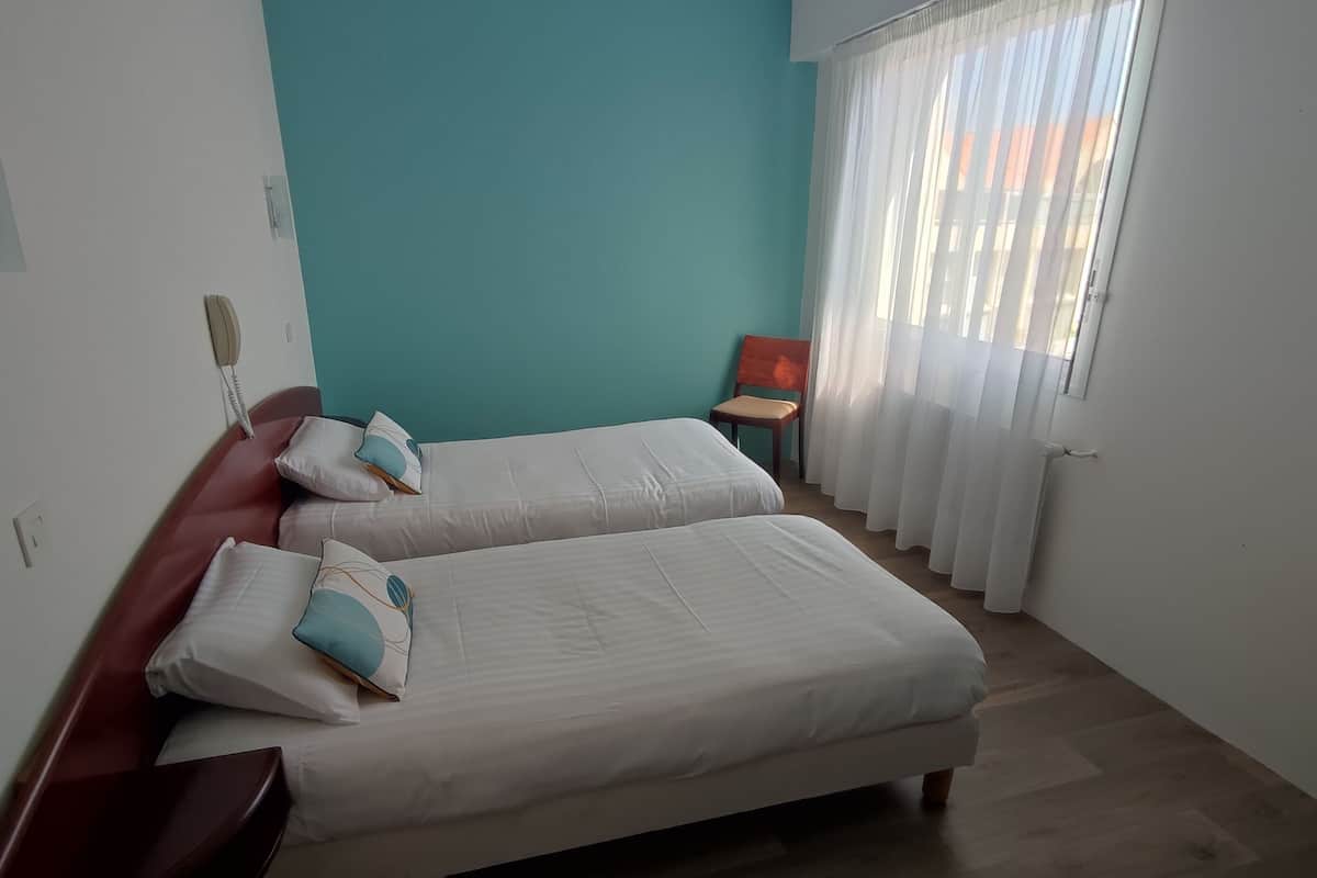 Ein Hotelzimmer mit zwei Betten, einem hölzernen Kopfteil, einem kleinen Tisch, einem Sessel und einem Fenster mit Vorhängen.