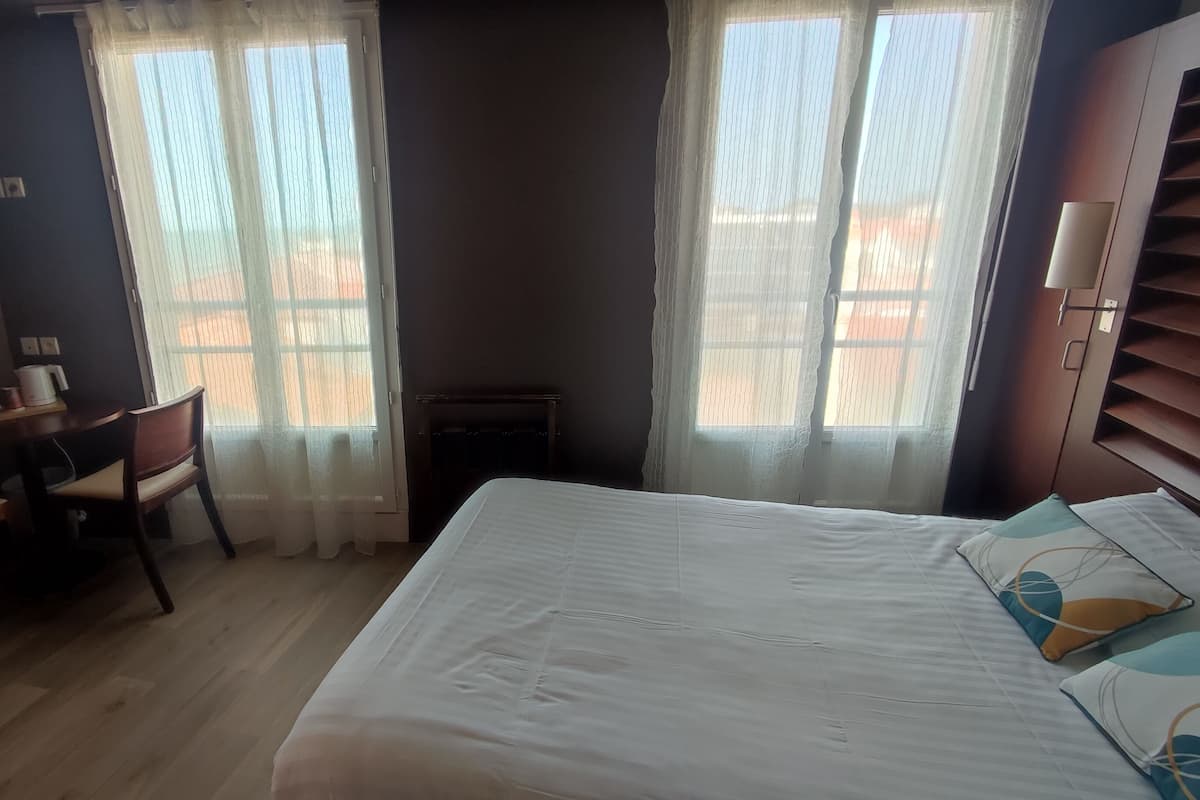 Ein Schlafzimmer mit einem Bett, einem Schreibtisch und zwei Fenstern mit durchsichtigen Vorhängen.