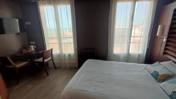 A bedroom with a bed, a desk, and two windows with sheer curtains.