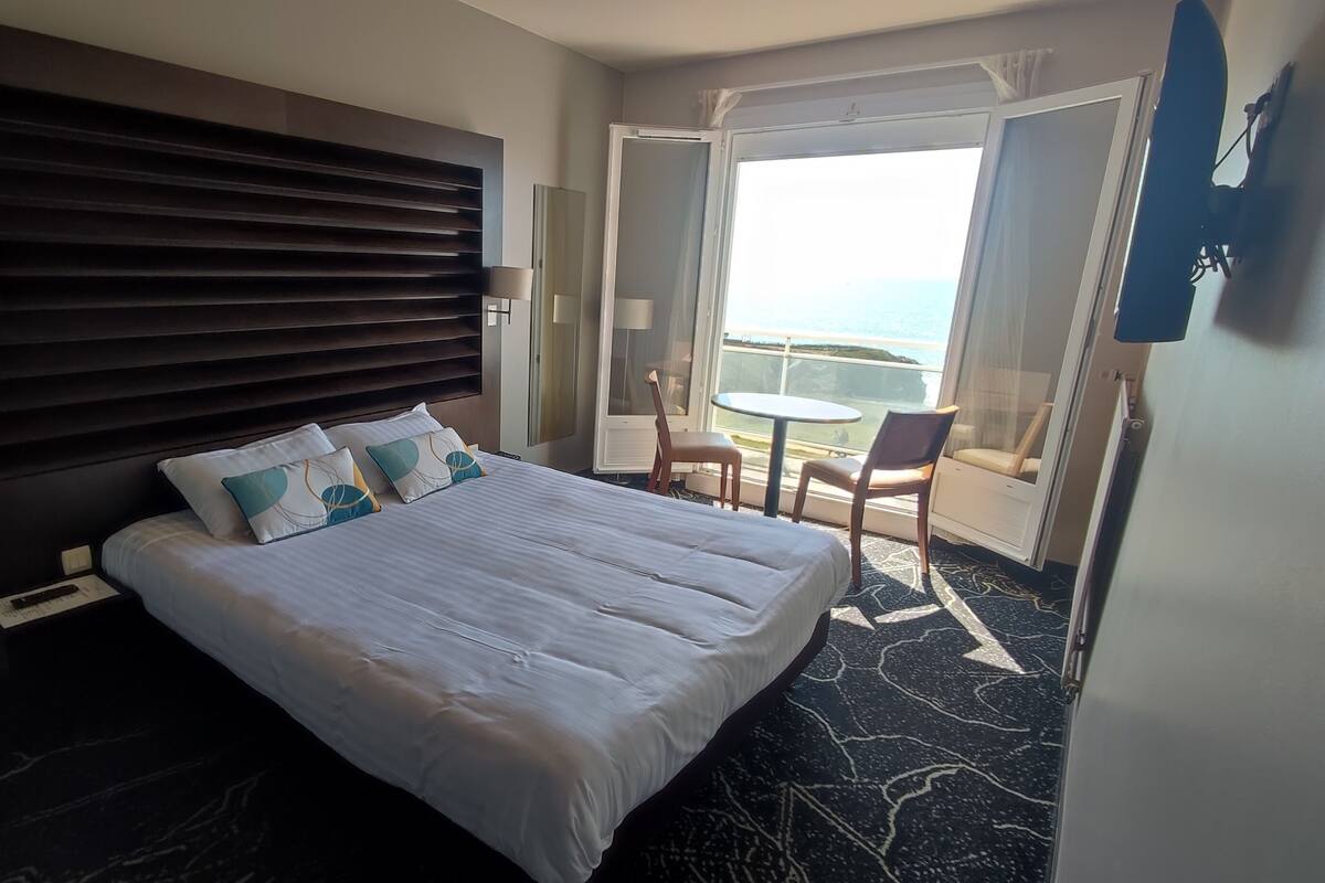 Ein Hotelzimmer mit einem großen Bett, einem Kopfteil, zwei Nachttischlampen, einem kleinen Tisch mit Stühlen und einem Balkon mit Blick.