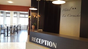 A hotel reception area with a reception desk labeled "RECEPTION," a wall with the hotel name "Hôtel La Corniche," and a seating area with tables and chairs.