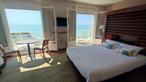 A hotel room with a large bed, a small table, a chair, and a balcony with a view of the sea.