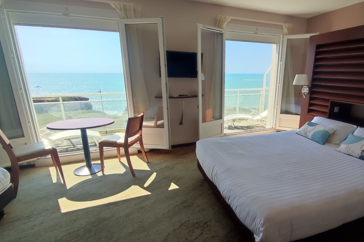 Ein Hotelzimmer mit einem großen Bett, einem kleinen Tisch, einem Stuhl und einem Balkon mit Meerblick.