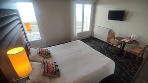 Quarto de hotel com cama, escrivaninha, cadeira, televisão e janela.