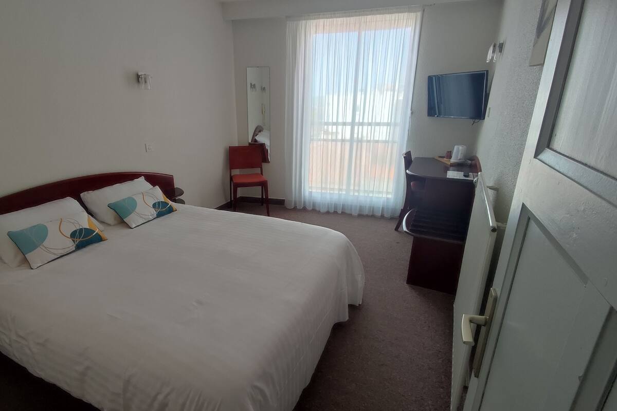 Ein Hotelzimmer mit einem Einzelbett, einem Schreibtisch mit Stuhl, einem Fernseher und einem Fenster mit Vorhängen.