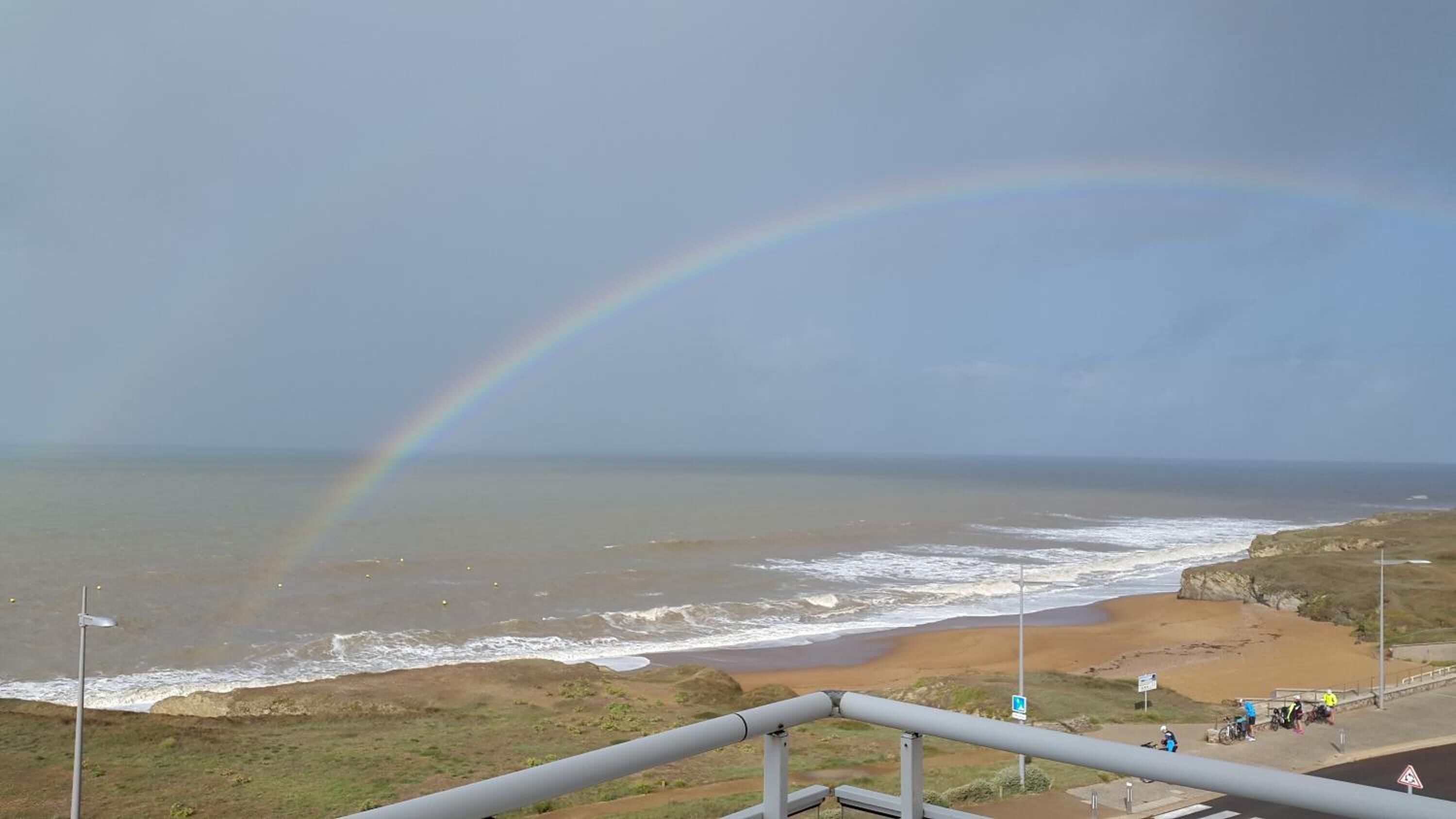 Vista costera con un arcoíris sobre el mar, una playa y una carretera con ciclistas.