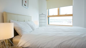 A neatly made bed with white linens in a room with a large window, a bedside table with a lamp, and a wall-mounted shelf. - Rethink Living - Hewitt Tower (Reading)