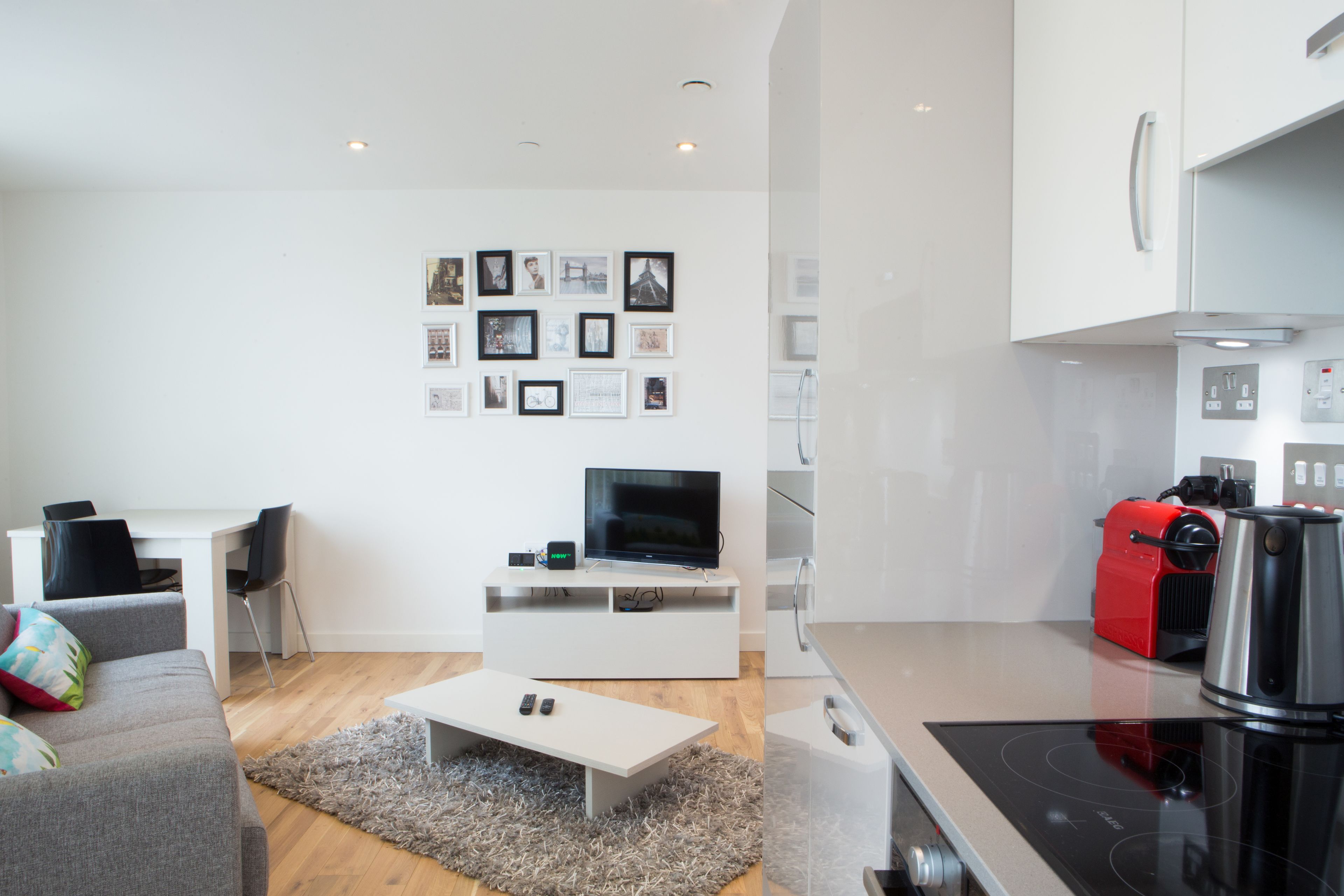 Una sala moderna con un sofá blanco, una mesa de centro y un televisor de pantalla plana. Se observa una zona de cocina con un electrodoméstico rojo y una estufa.