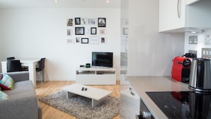 A modern living room with a white sofa, a coffee table, and a flat-screen TV. There is a kitchen area with a red appliance and a stove. - Rethink Living - Hewitt Tower (Reading)