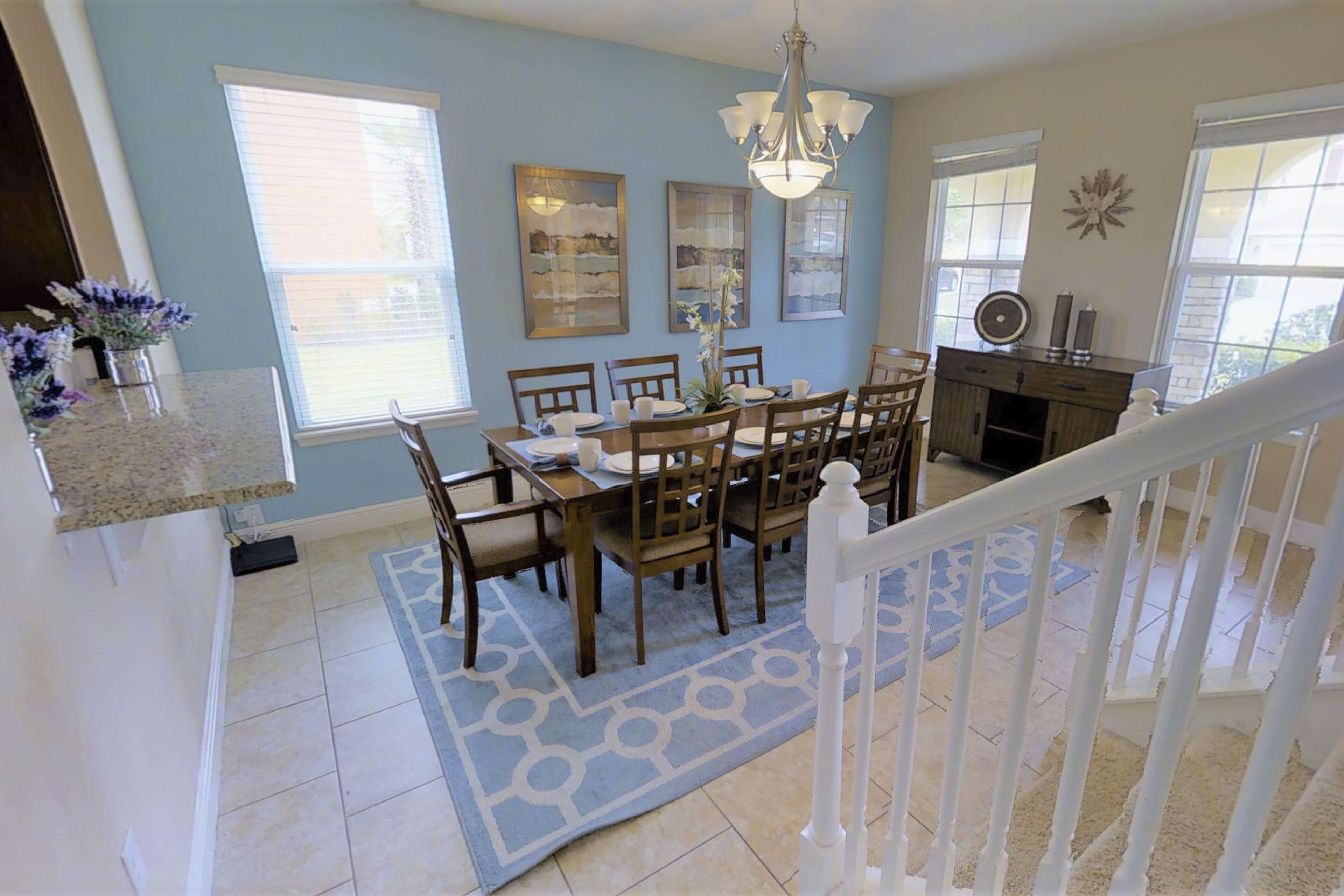 A dining area with a table set for six, surrounded by wooden chairs, a blue patterned rug, and a chandelier.