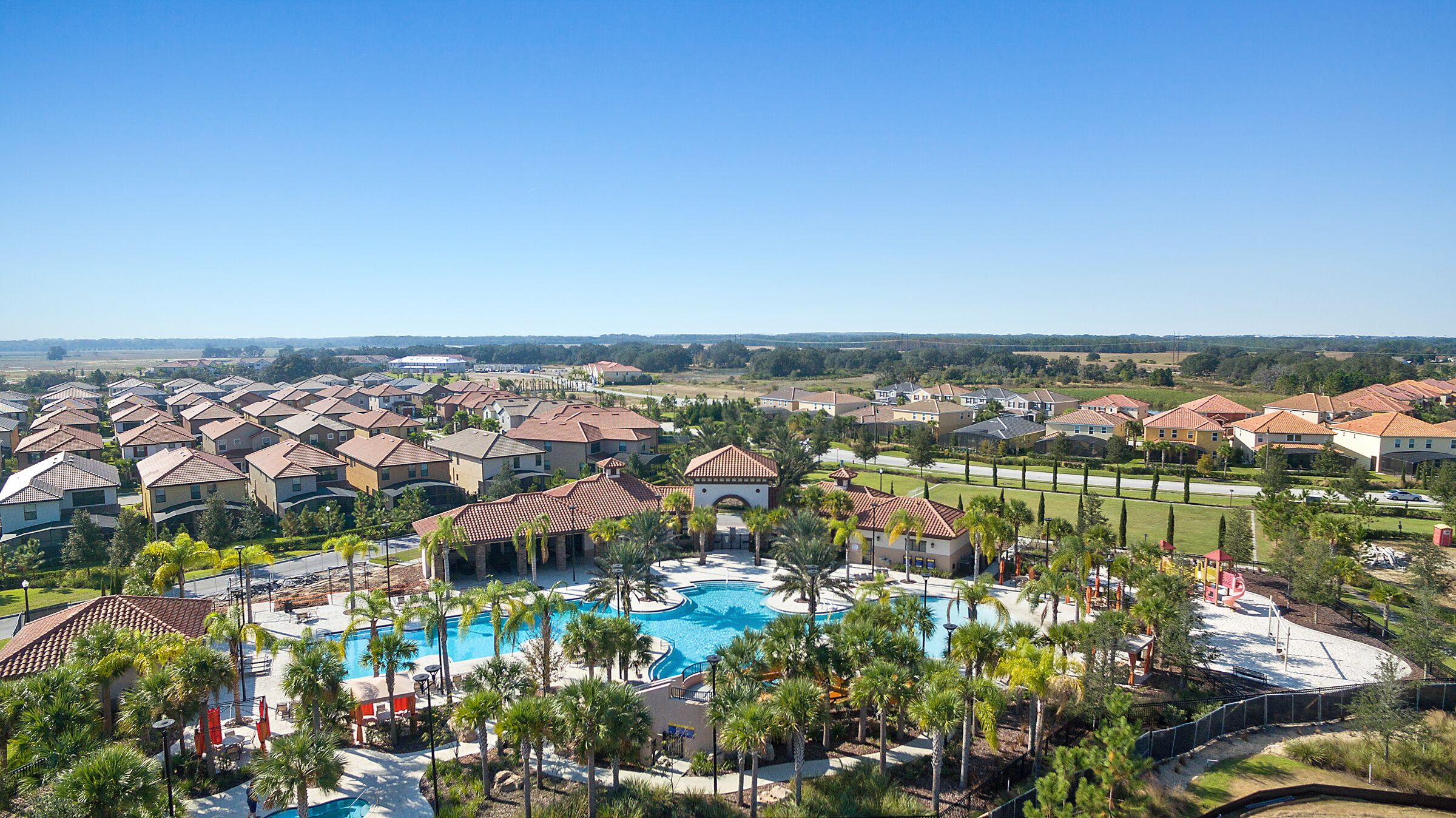 A residential area with a central swimming pool surrounded by palm trees and houses with red-tiled roofs.