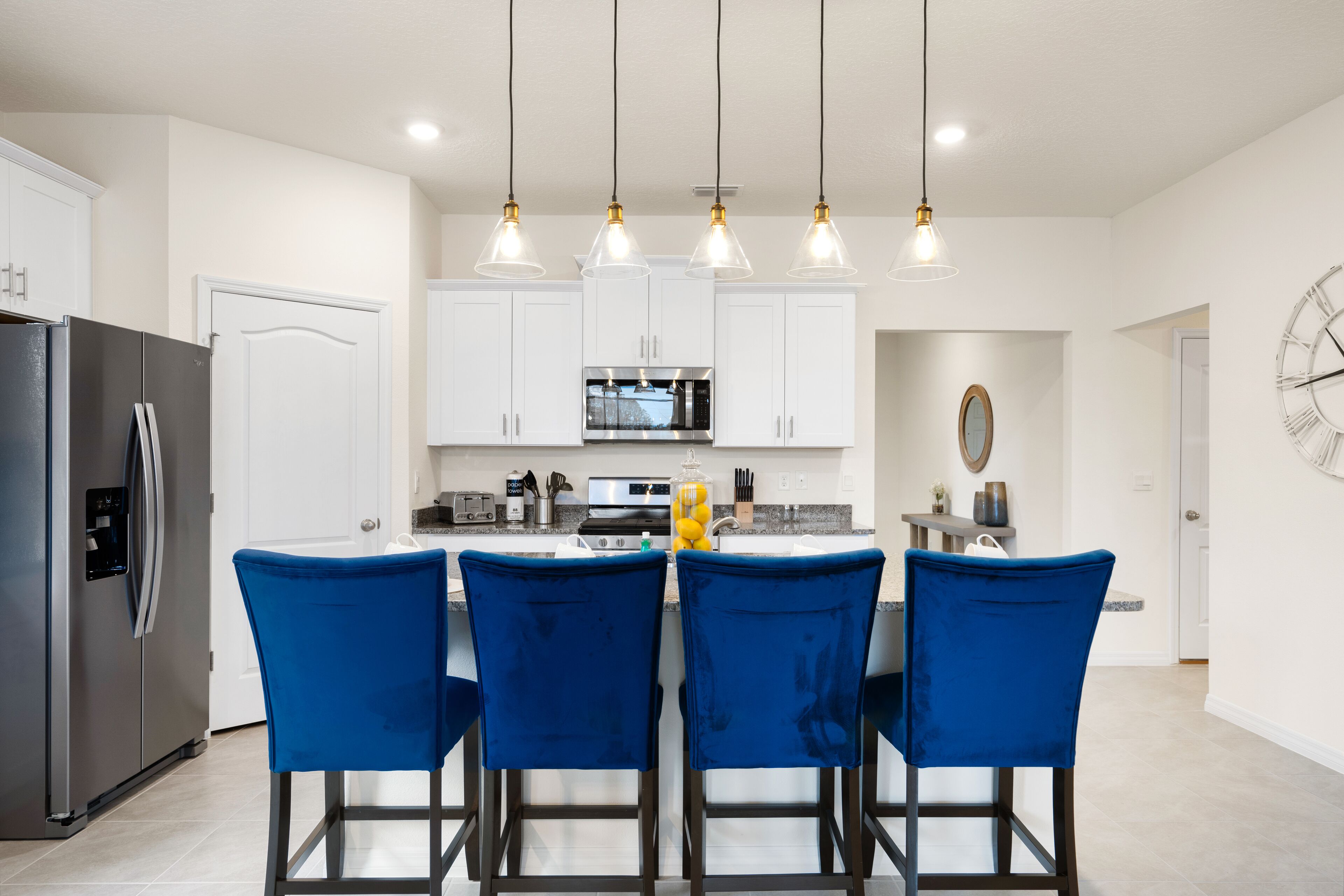 A modern kitchen with white cabinets, a stainless steel refrigerator, a microwave, and a sink. There are four blue chairs at the kitchen island.
