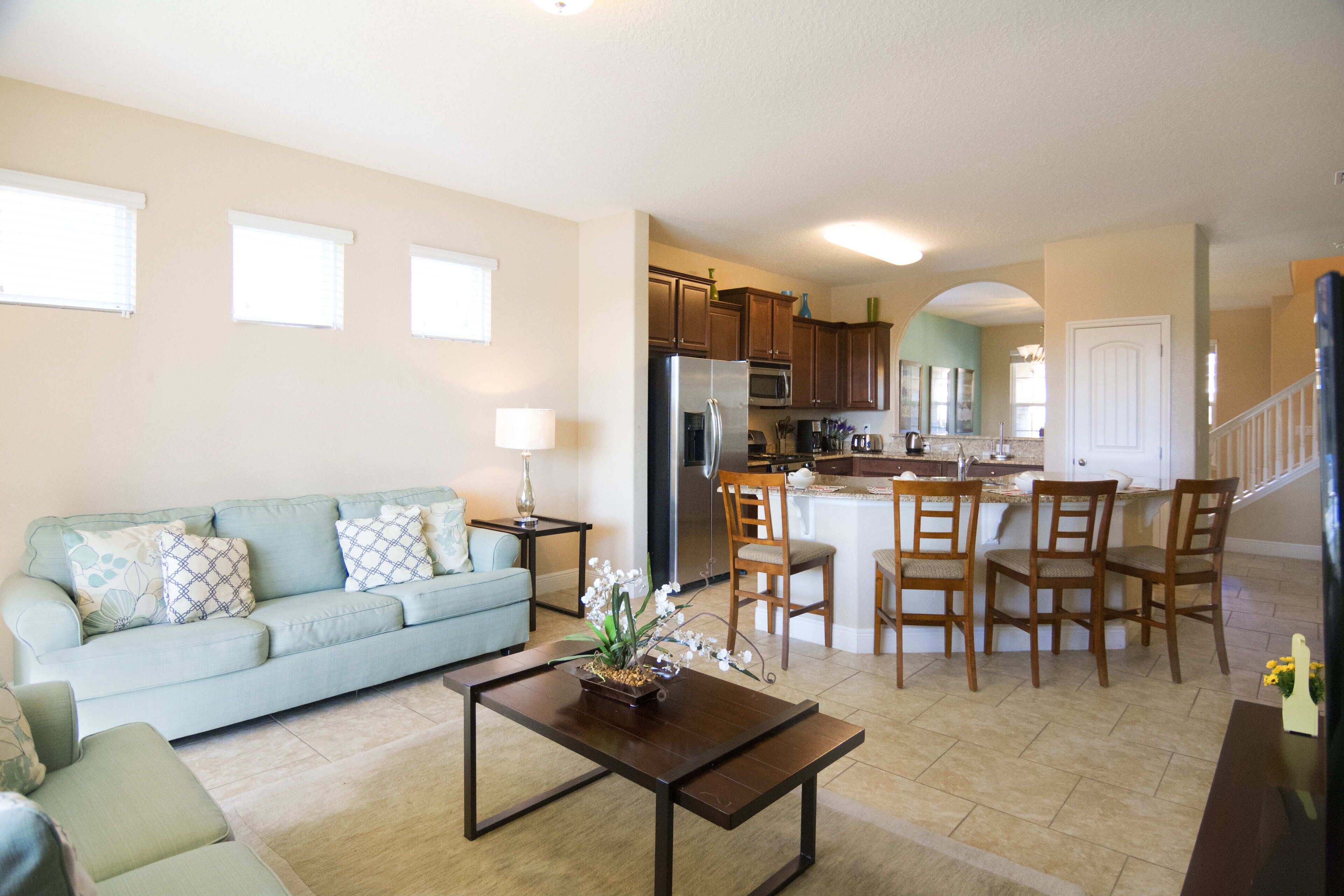 A spacious living room with a sofa, coffee table, and a kitchen area with a refrigerator, microwave, and dining chairs.