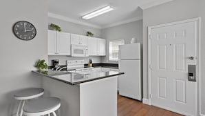 A modern kitchen with white cabinetry, a central island, two bar stools, a microwave, a refrigerator, and a clock on the wall. - Lucaya Vacation Homes (Kissimmee)