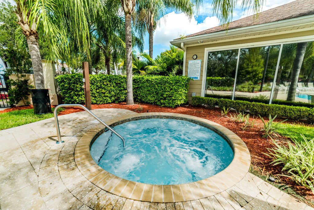 A hot tub surrounded by a landscaped garden with palm trees and a building with a pool in the background.