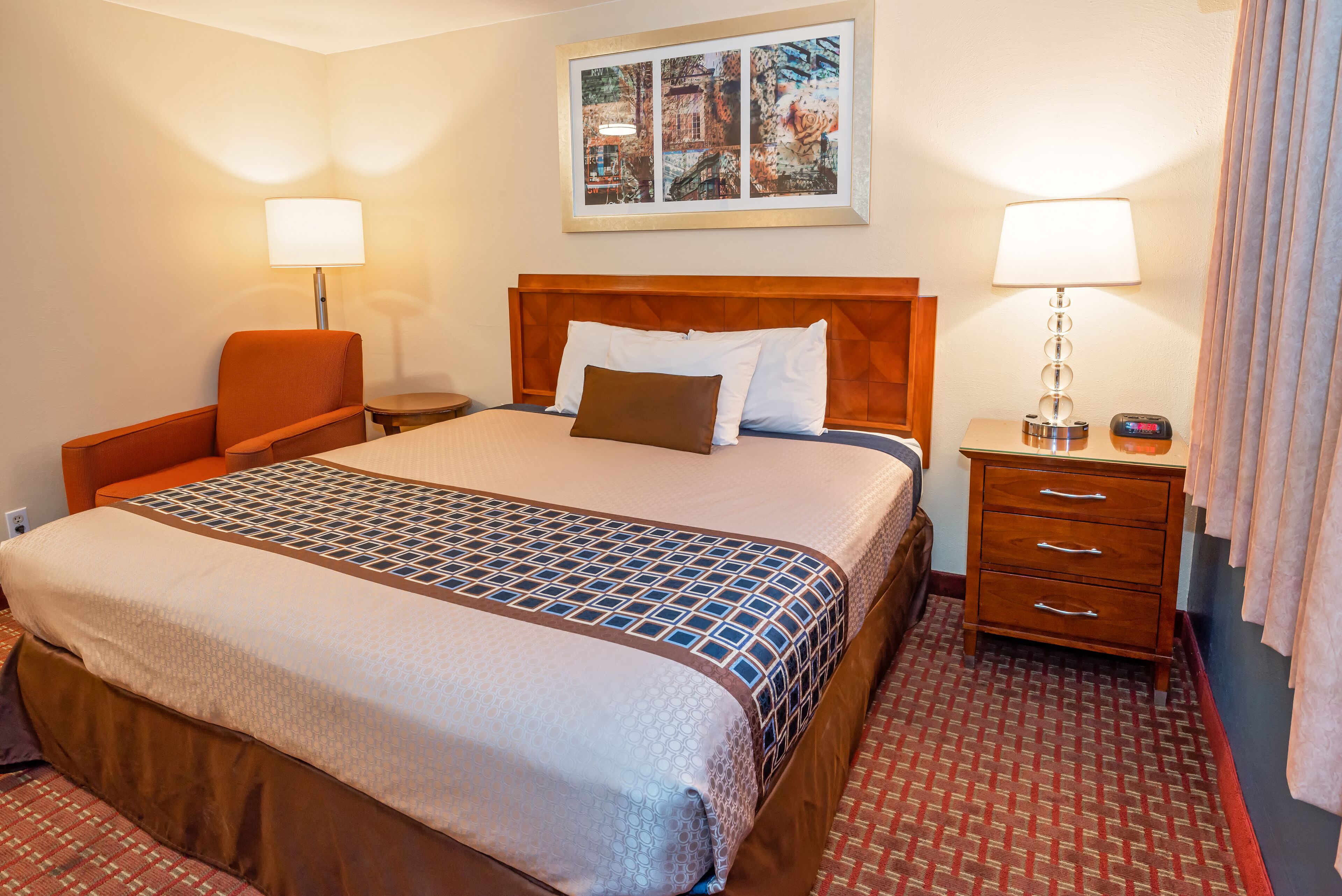 A hotel room with a large bed, a wooden headboard, two bedside tables with lamps, a chair, and a red patterned carpet.