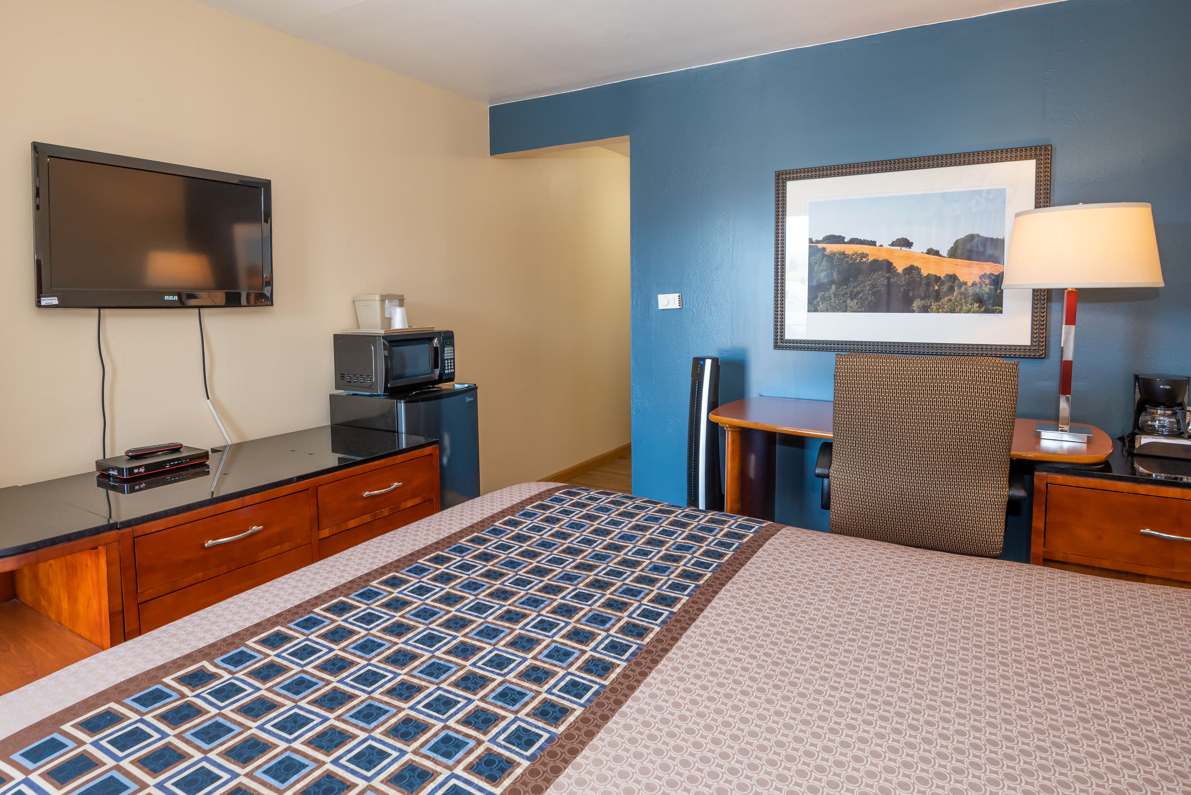 Ein Hotelzimmer mit einem Bett, einem Flachbildfernseher, einer Mikrowelle, einem Schreibtisch, einem Sessel und einem gerahmten Bild an der Wand.
