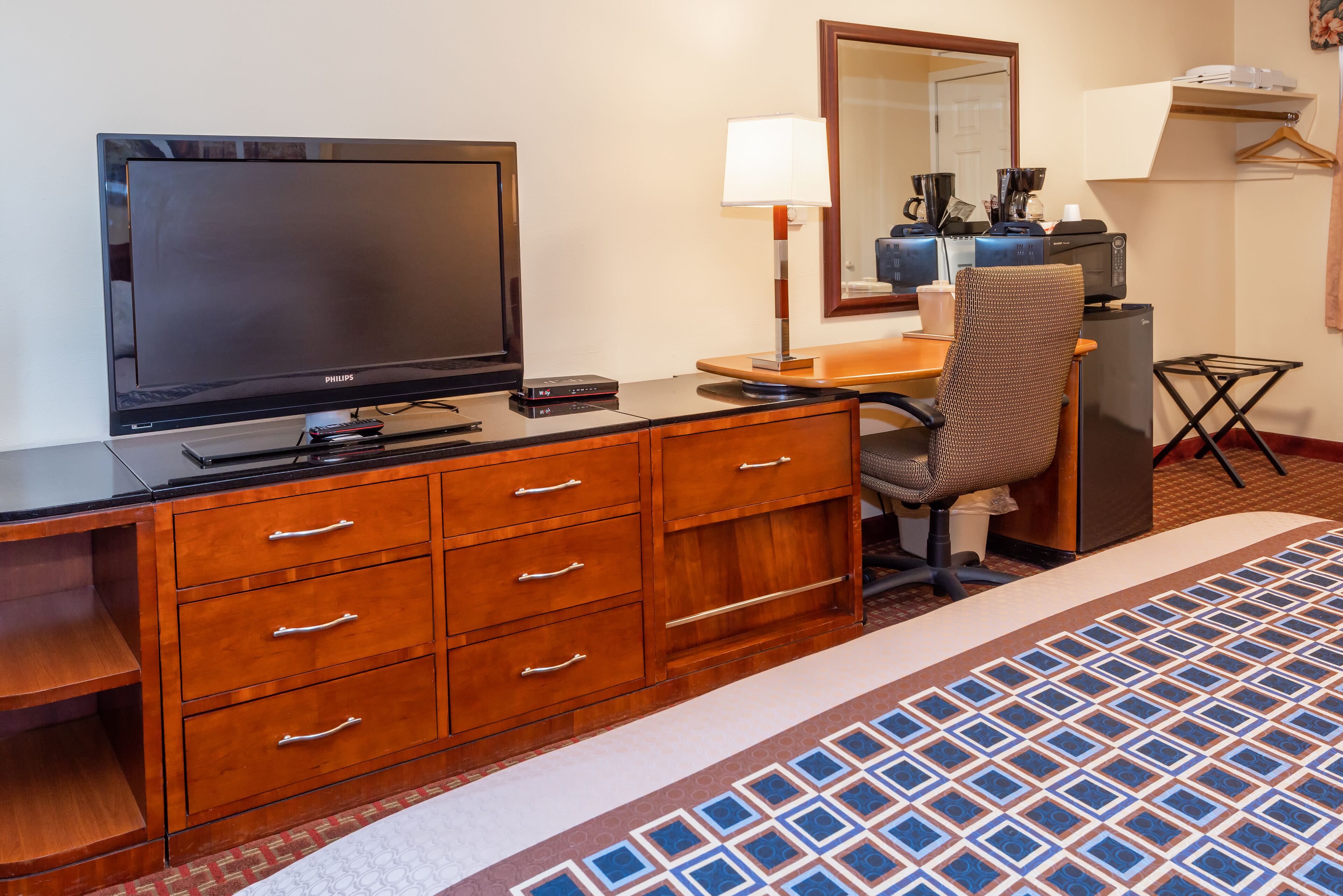 A hotel room with a wooden dresser, a television, a desk with a lamp, a chair, a small table, and a patterned rug.