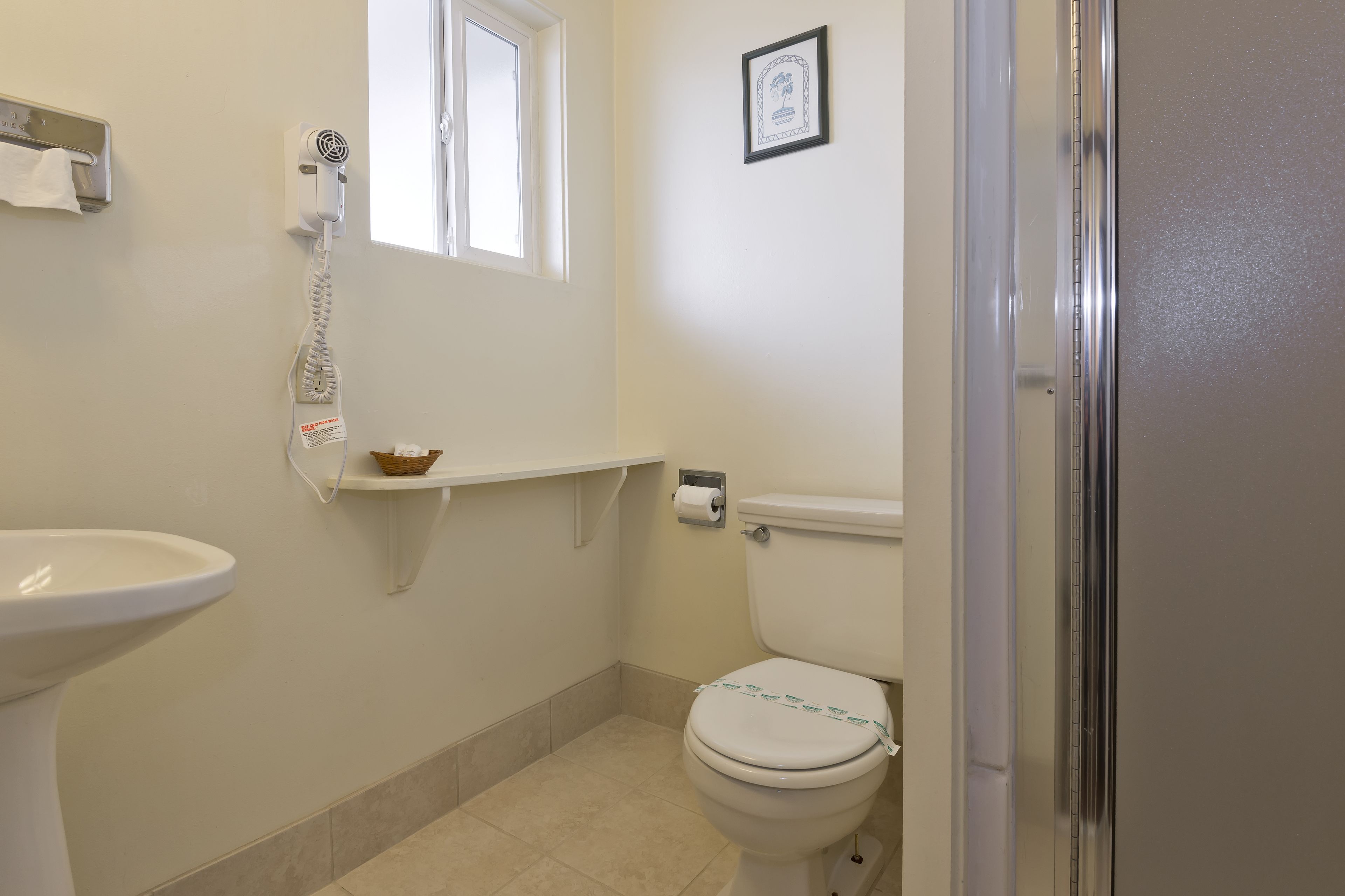 A bathroom with a toilet, a sink, a window, a framed picture on the wall, and a towel dispenser.