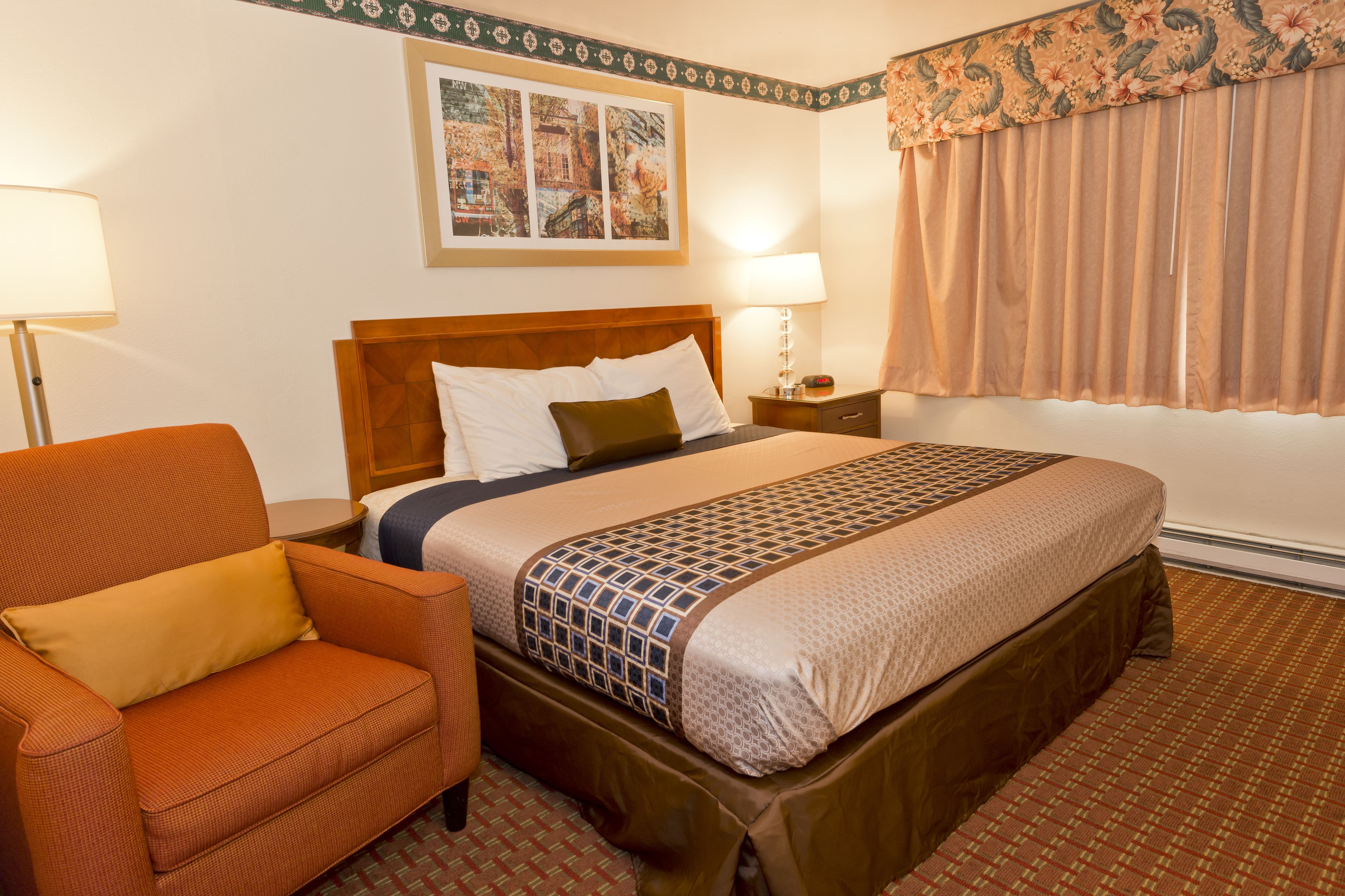A hotel room with a bed, a chair, a nightstand, and a wall with artwork.