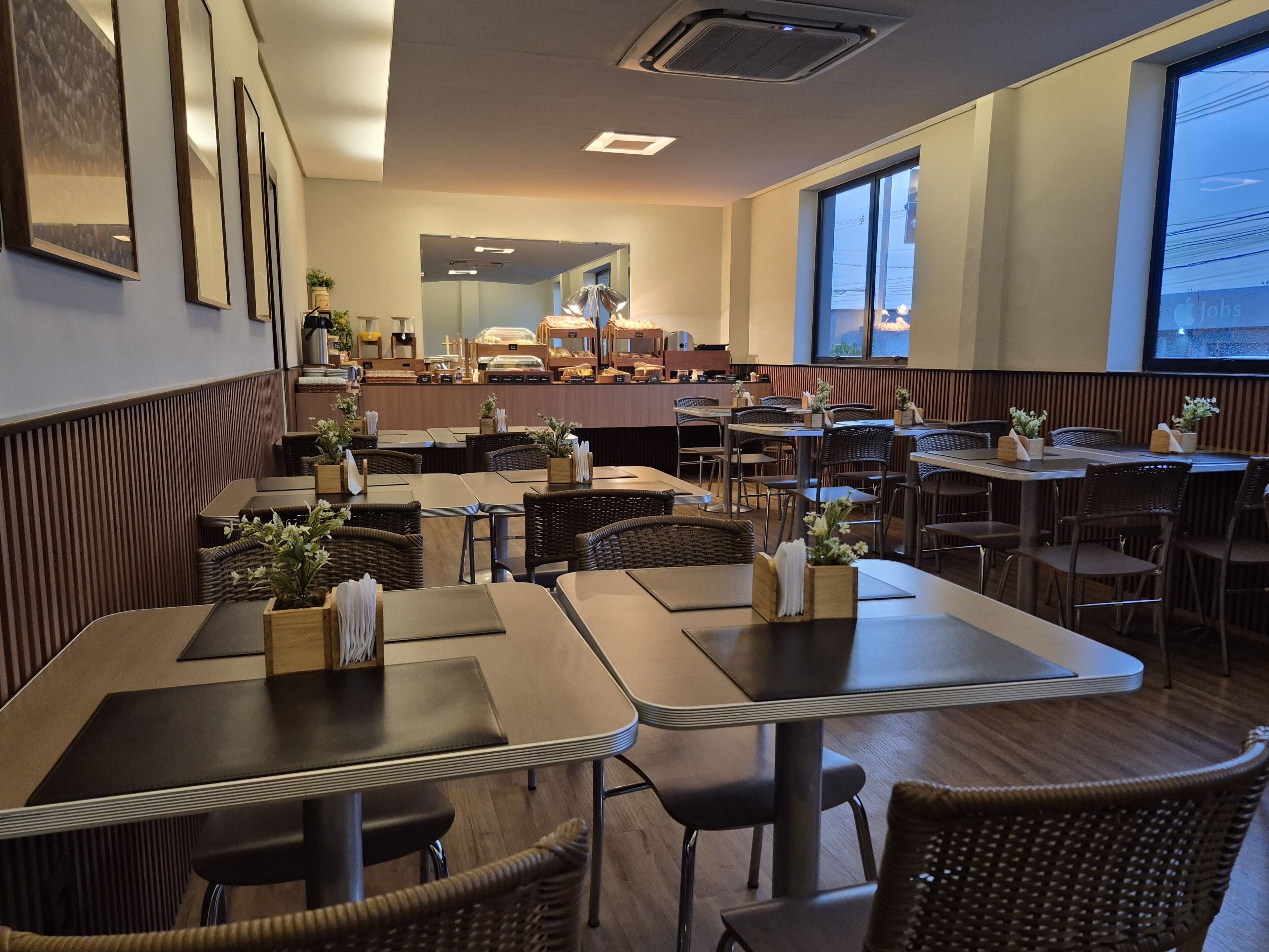 Interior de restaurante con varias mesas y sillas, una barra con vitrina y grandes ventanales que dejan entrar luz natural.