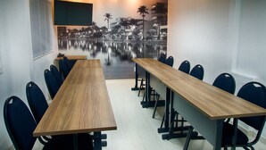 A conference room with a long wooden table and multiple chairs, a large flat-screen TV on the wall, and a mural of a cityscape with palm trees. - Real Hotel (Sete Lagoas)