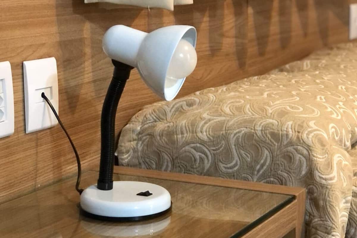 Uma mesa de cabeceira com abajur branco, tomada e cama com roupa de cama estampada.