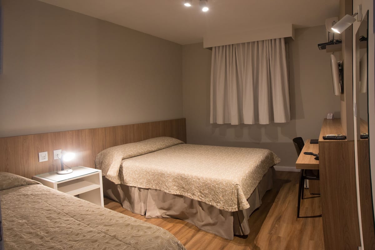 Quarto de hotel com duas camas, uma escrivaninha com cadeira, luminária, televisão de tela plana e janela com cortinas.