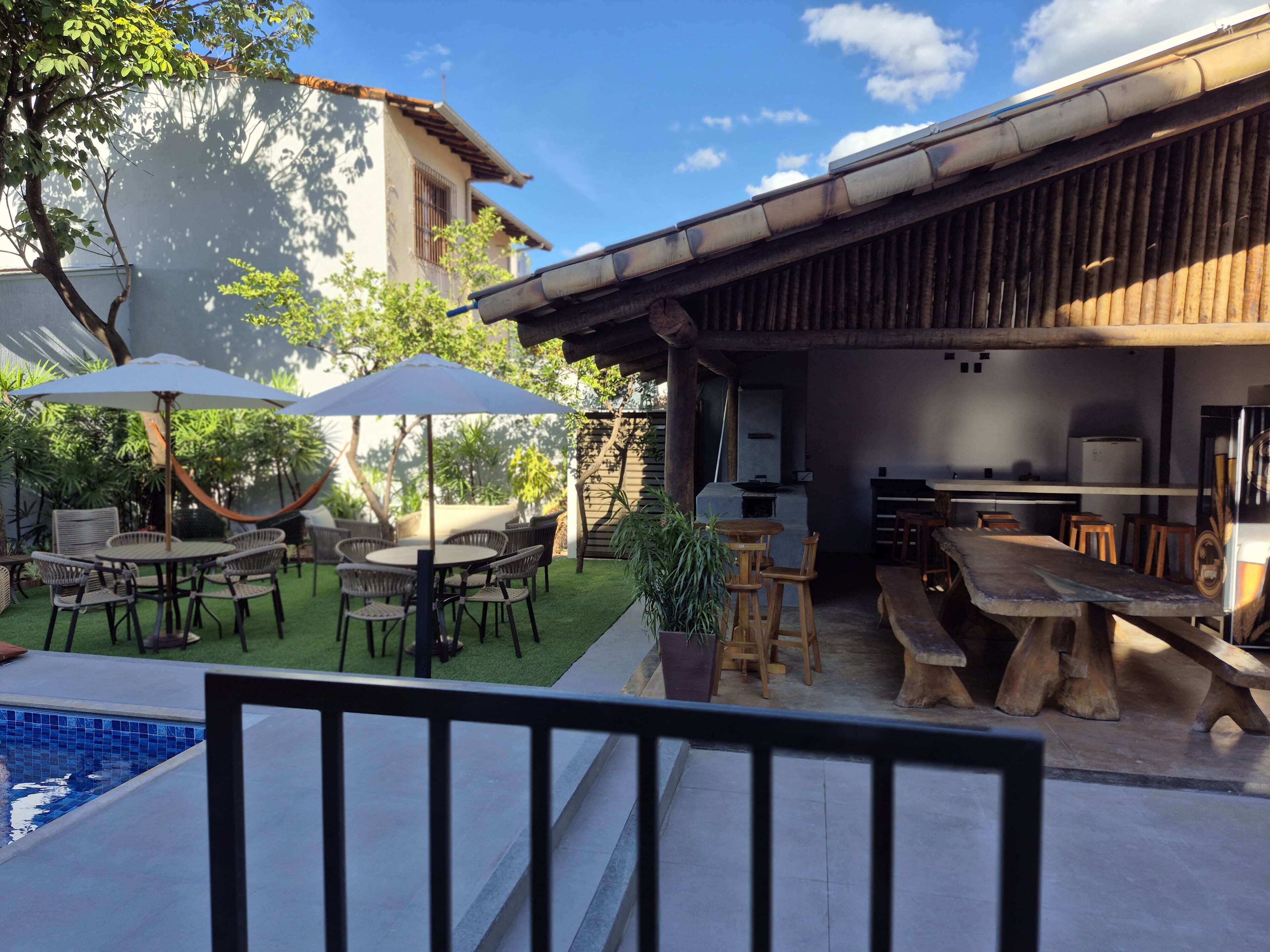 Uma área de pátio externa com mesas e cadeiras redondas, uma piscina e um bar com banquetes.