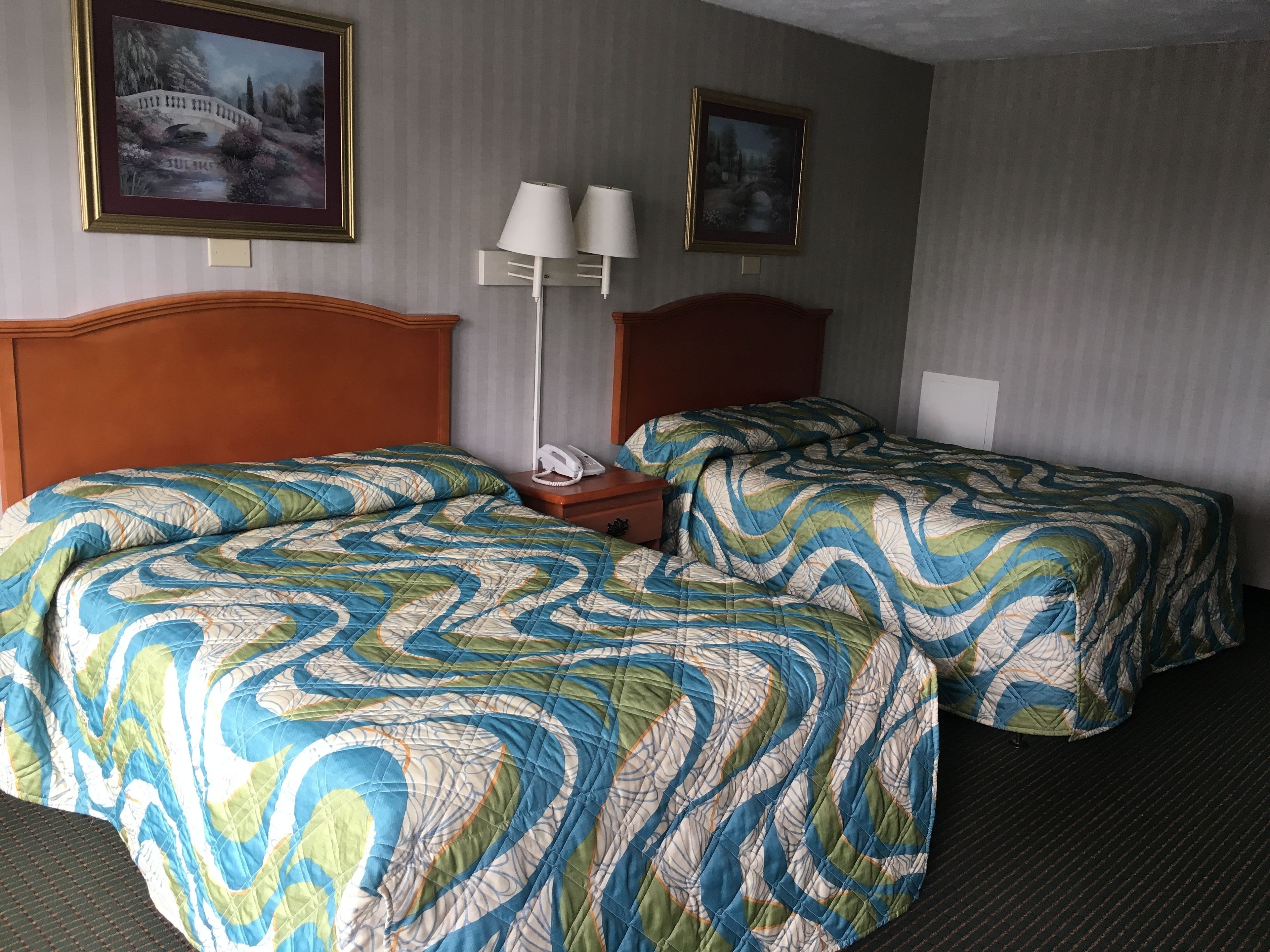 A hotel room with two beds, each with a patterned bedspread, a wooden headboard, a bedside table with a lamp, and two framed pictures on the wall.
