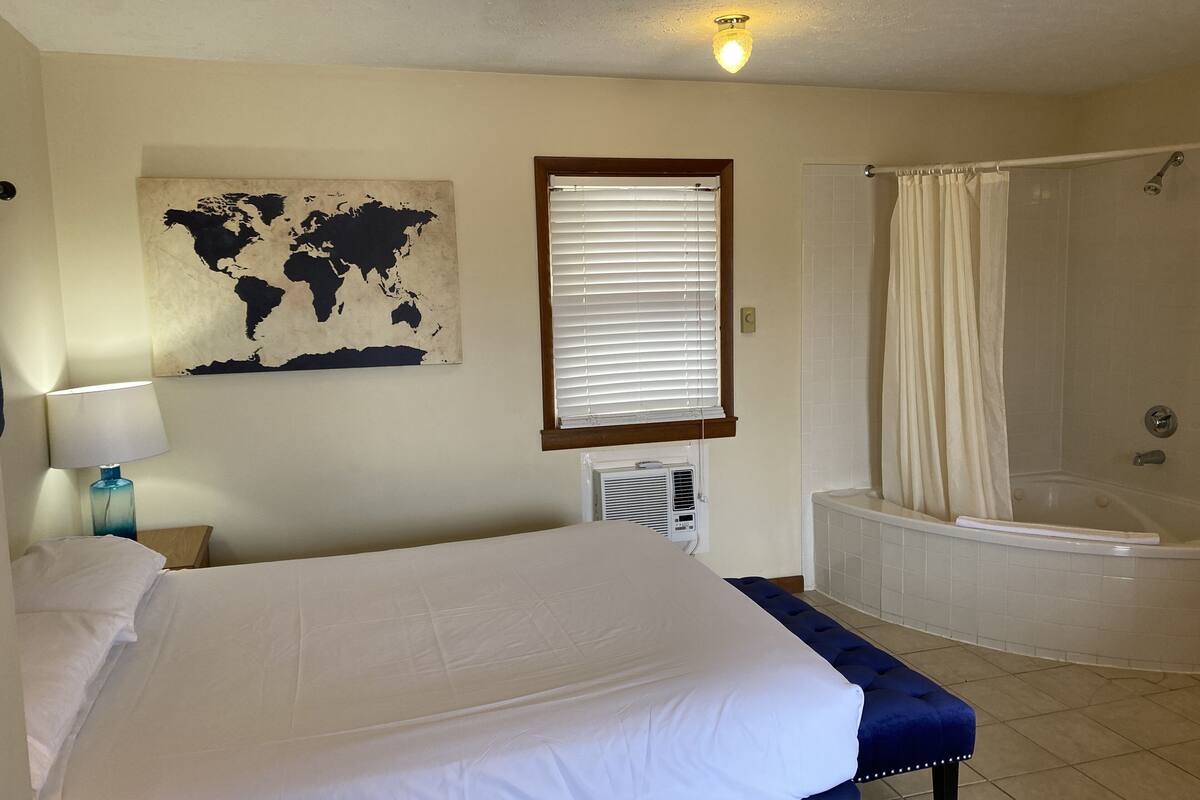 A hotel room with a large bed, a world map artwork, a window with blinds, an air conditioning unit, and a separate bathroom with a bathtub and shower.
