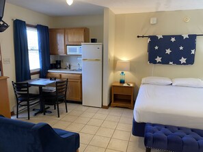 A compact hotel room with a kitchenette, a bed with a star-patterned headboard, a desk with a chair, and a small bedside table with a lamp.
