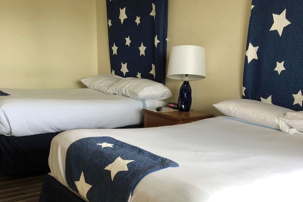 A hotel room with two beds, a nightstand with a lamp, and star-patterned curtains.
