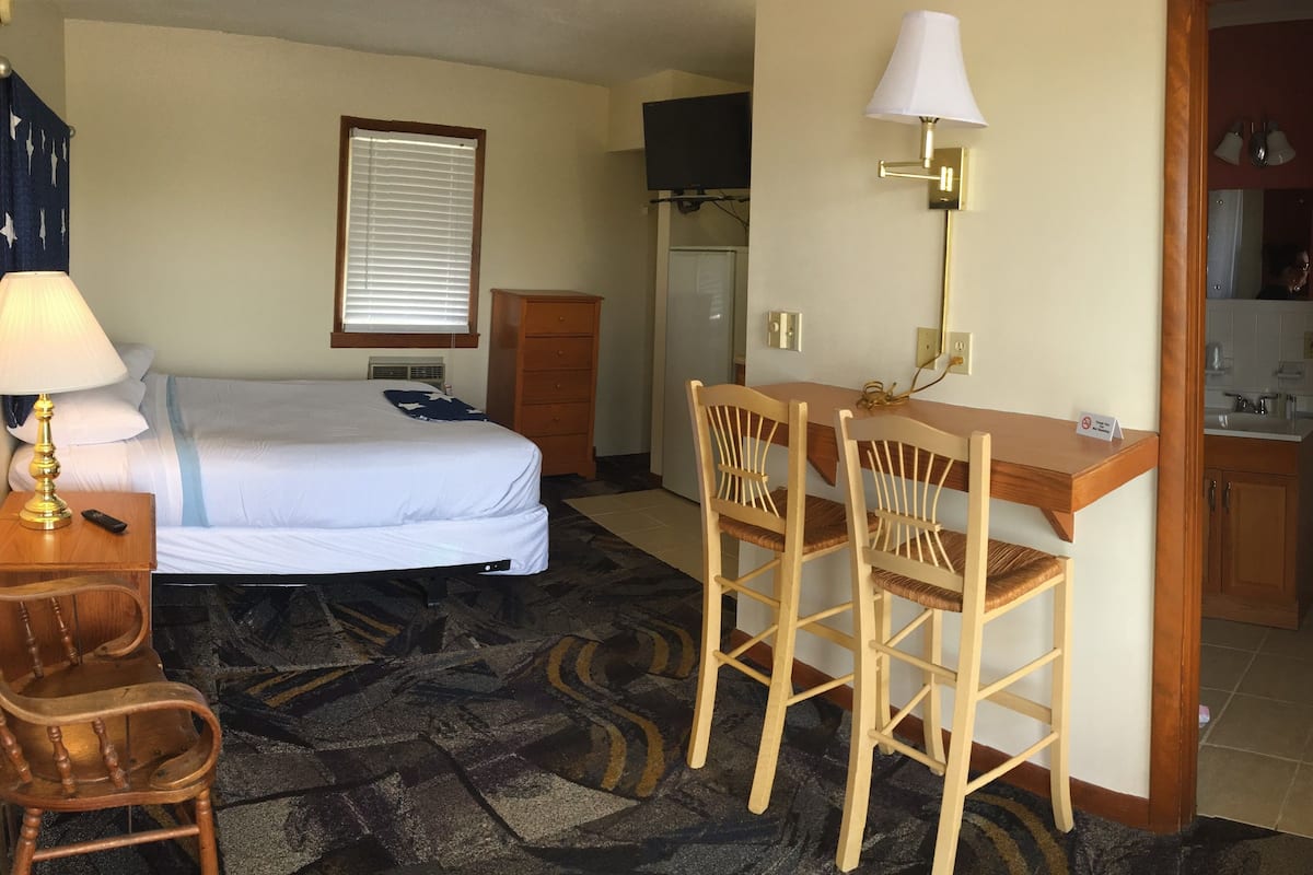 A hotel room with a bed, desk, two chairs, a lamp, a dresser, a television, and a bathroom visible through an open door.